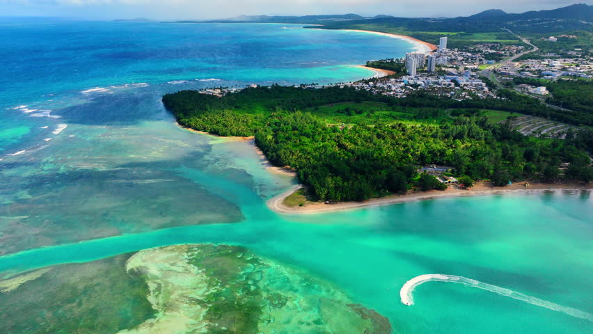 Coastal town featuring white buildings, dense green forest, sandy beaches, coral reefs, and a jet ski trail in clear blue ocean water. Aerial view of tropical coastline Puerto Rico.