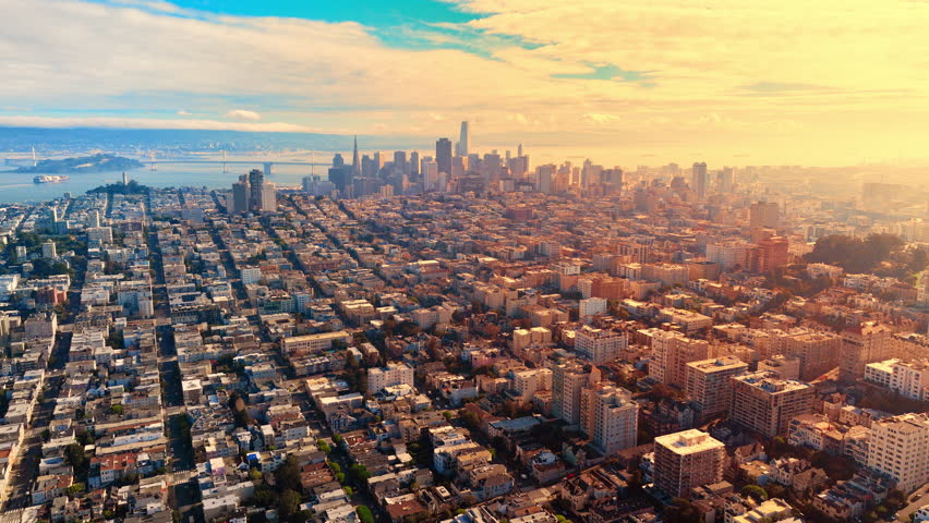 City blocks, residential buildings, and streets leading towards the distant financial district under a warm sunset sky. Dense urban landscape of San Francisco residential area at sunset.