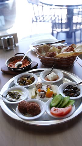 Close view of a traditional Turkish breakfast spread served on a metal tray with olives, cheese, vegetables, yogurt dip, bread basket, and small dishes on a restaurant table in natural daylight.