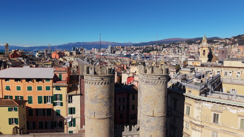 Drone ascends in front of Porta Soprana towers in Genoa, Italy, revealing the dense historic center, colorful buildings, harbor area and Ligurian mountains under clear sky.