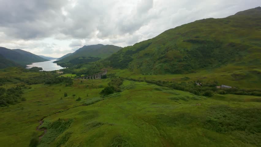 Aerial view over the Glenfinnan Viaduct, banking right to showcase the iconic stone arches with the expansive, deep blue waters of Loch Shiel serving as a majestic backdrop in the Scottish Highlands.