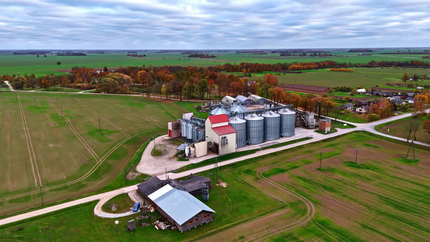 Aerial parallax shot of grain storage silos and elevator facility surrounded by green farmland and autumn trees in rural countryside