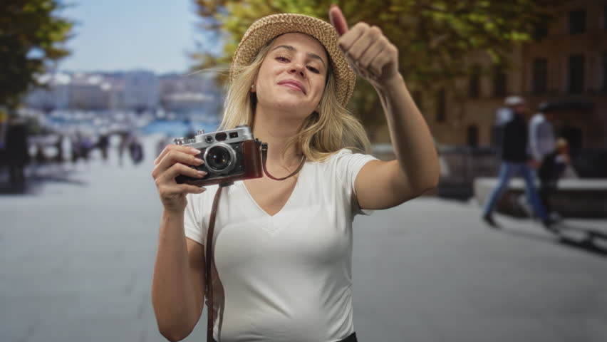 Woman holding vintage camera and showing thumbs up on street by trees and stone benches, wearing straw hat and white tshirt; travel joy.