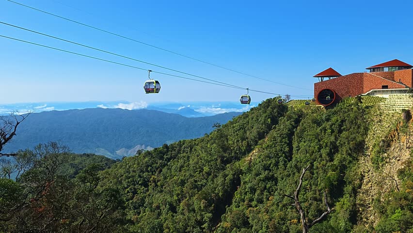 Cable car gondolas arriving at scenic Ba Na Hills mountain peak station with red-brick building, vast cloud-filled valley and blue mountain ranges beyond