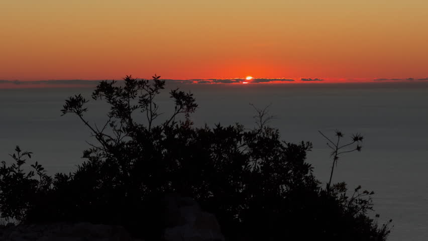 Dramatic red sunset over a calm sea, seen from a cliff with dark foliage silhouettes and vivid sky reflections glowing on the water.