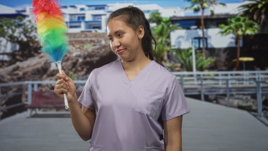 Woman cleaner in scrub top holding rainbow feather duster and giving thumbs up on an outdoor pier in front of a building; pride.