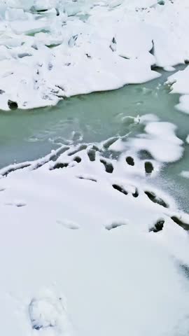 Boot prints in fresh snow beside a thin ice sheet and dark meltwater gaps, creating a minimal winter texture and high-contrast abstract shoreline scene with copy space, vertical.