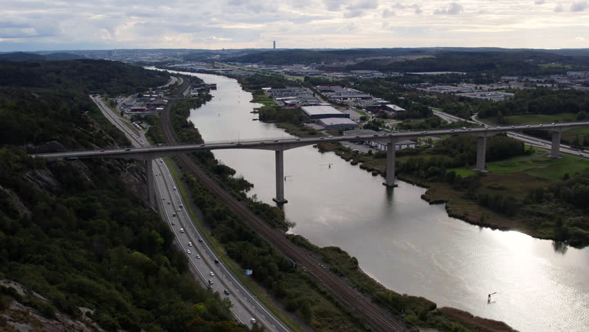 Drone shot of Angeredsbron bridge spanning a wide river in Gothenburg, Sweden. A busy highway and railway run parallel below, with an industrial cityscape and green hills stretching to the horizon.