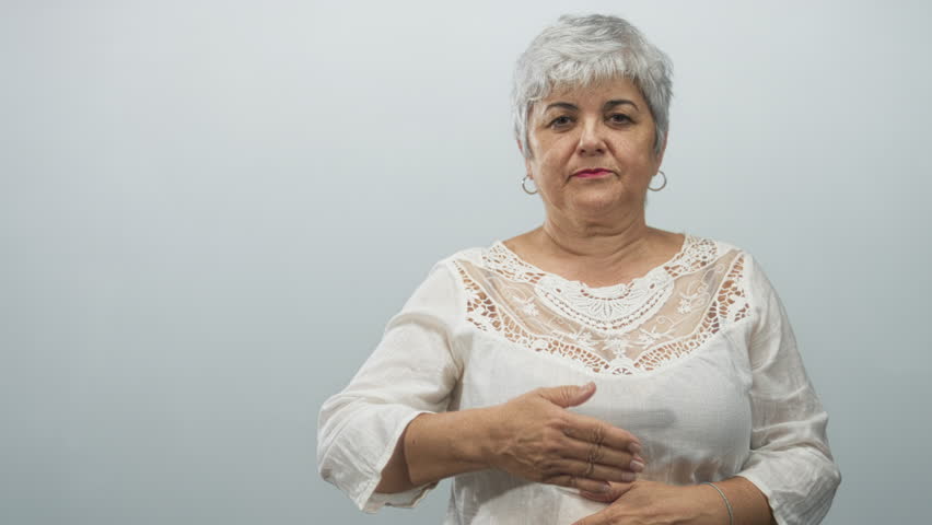 Woman with grey hair and hoop earrings smiling raises right hand to cover one eye while left hand rests on stomach in studio with plain backdrop; playful joy.