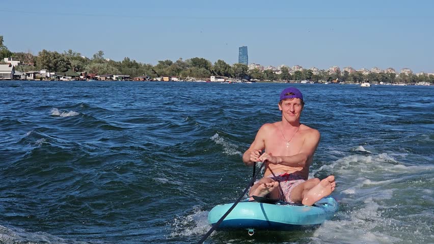 Man is sitting on a surfboard in the ocean. He is wearing a purple bandana and is holding a surfboard leash