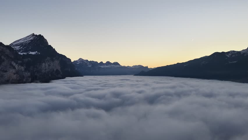 Soft sunrise light spreads across a vast sea of fog covering Walensee valley in the Swiss Alps. Peaceful alpine winter scene with dramatic silhouettes and tranquil morning atmosphere.