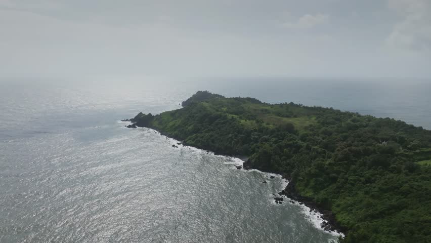 Drone footage glides over a lush green peninsula with steep cliffs descending into ocean waters, white waves crashing against dark rocky shoreline under overcast skies.