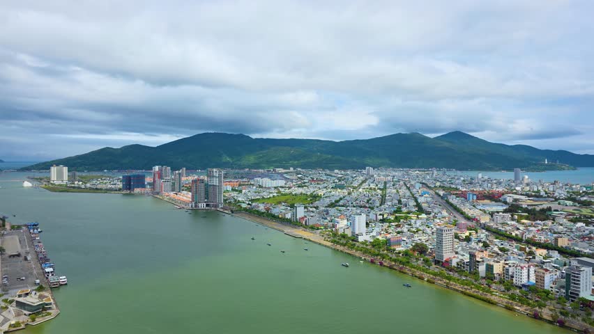 Zoom-in hyperlapse of Da Nang cityscape along Han River with dense residential districts, waterfront development and Son Tra mountain under dramatic moving clouds