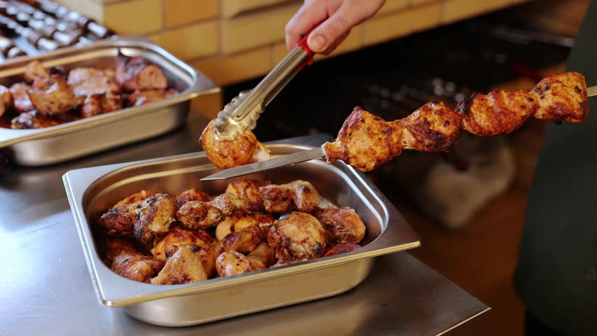 Man hand using metal tongs to transfer hot, grilled shish kebab from skewer to tray