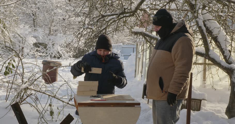 A caring father teaches his little son how to build a wooden bird feeder in his snowy backyard. Father and son spend time together and help wildlife by creating a bird feeder on a cold winter day.
