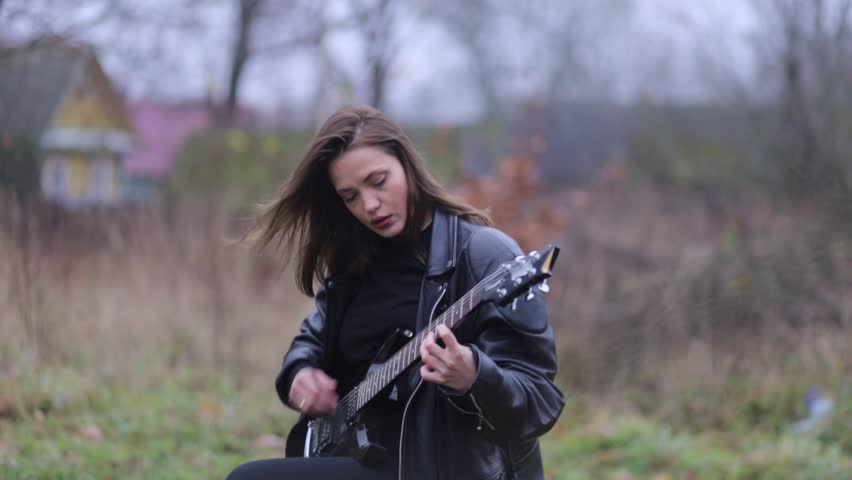 Young woman in a leather jacket, rock musician plays electric guitar outdoors