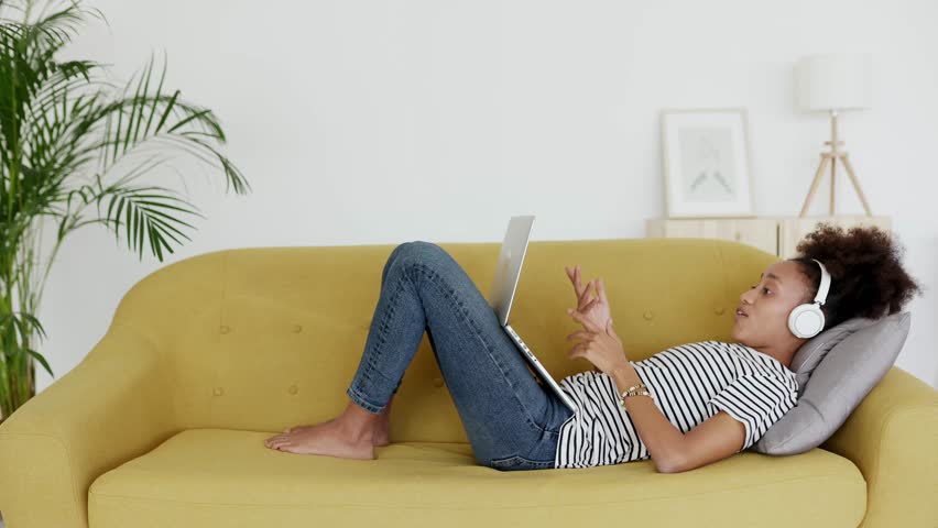 Cheerful young African woman with headphones lying on a comfortable yellow sofa at home, happily video calling, laughing and talking with friends or family using a modern laptop computer