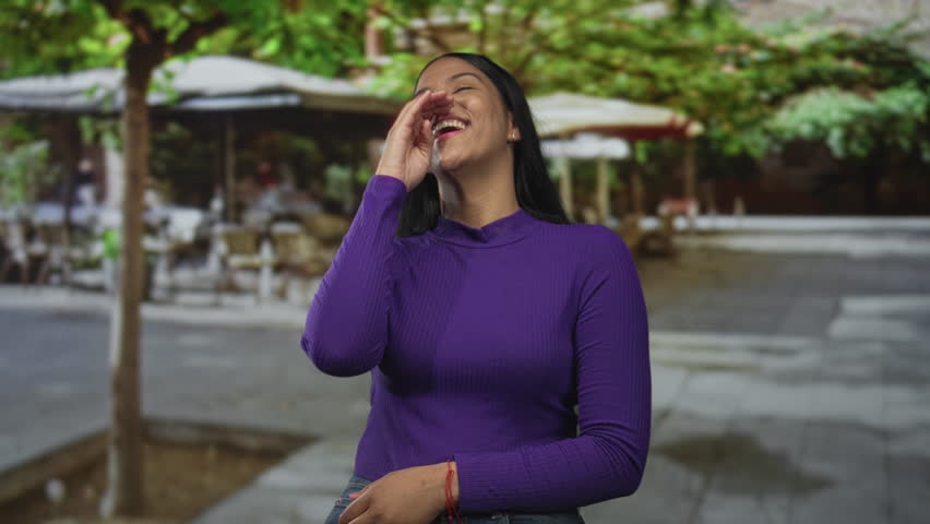 Woman in a purple top covering her mouth with hand while laughing on a leafy city street under cafe umbrellas; happiness.
