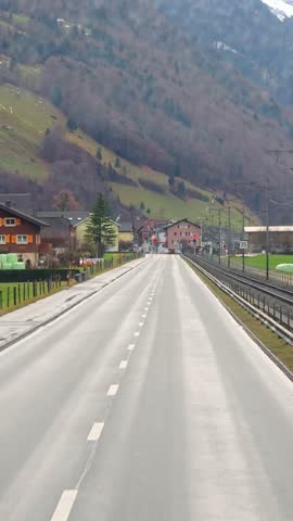 Rural Swiss village road beside railway tracks with alpine hills and houses in scenic mountain landscape.
