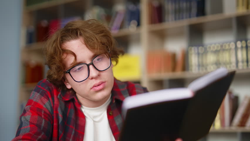 Young male student with curly hair wearing eyeglasses sits at a desk in a library, attentively reading a textbook and moving his lips as he studies the material with a focused expression