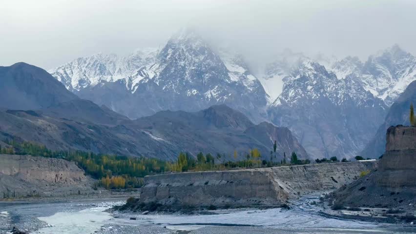 This is a horizontal video of snow-covered mountains with a river flowing below in northern Pakistan. It is foggy and cloudy, with alternating green and yellow poplar trees in the background.