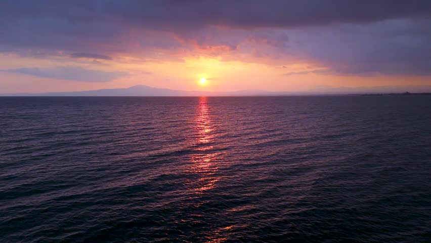Aerial drone footage of sunset over the Mediterranean Sea in Greece, with calm water, bright sun reflection, distant mountains on the horizon, and warm evening sky with soft clouds.
