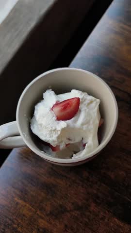Close up top view of a person eating a delicious strawberry and cream dessert. A spoon scoops the creamy treat from a mug on a wooden table