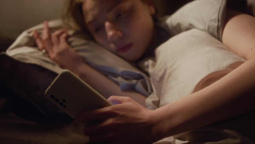 Young adult woman reading news on phone in bed after waking up in morning then turning back to sleep feeling overwhelmed