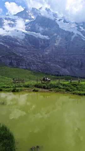 Majestic snow‑capped mountains in Switzerland rising above green alpine meadows. A couple sitting on a bench overlooking the mountains.