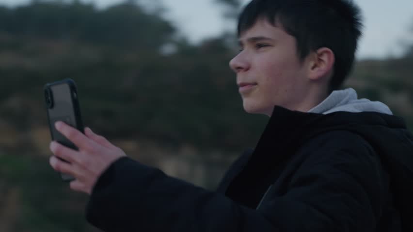 A happy teenage boy takes a photo with his smartphone while on a trip in nature. He is smiling and enjoying the scenery.
