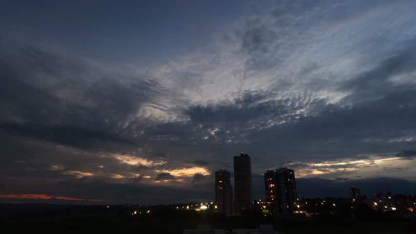 Dramatic blue hour sky with altocumulus clouds over a Brazilian city skyline at dusk, capturing the transition from day to night. - A stunning wide-angle shot capturing the magical blue hour over a modern Brazilian city. The deep, vibrant blue sky is filled with beautifully textured altocumulus clouds, creating a dramatic and moody atmosphere. Below, the city
