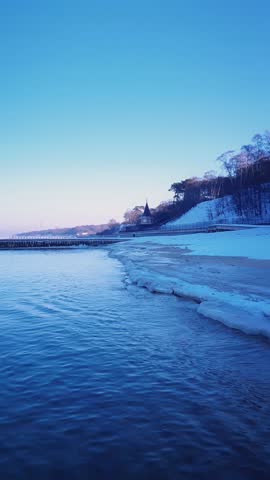 Vertical shot tranquil seaside landscape with calm coastal surf water waves rolling on wood breakwater and frozen snowy shore on Baltic sea coast under clear blue sky at frosty winter morning or dusk