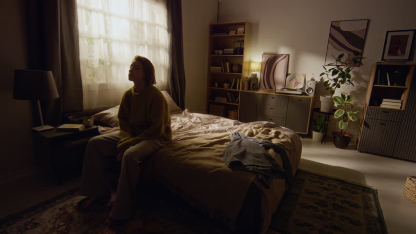 Wide angle shot of sunlit bedroom with young adult woman holding head coping with anxious thoughts then falling onto bed feeling fatigued
