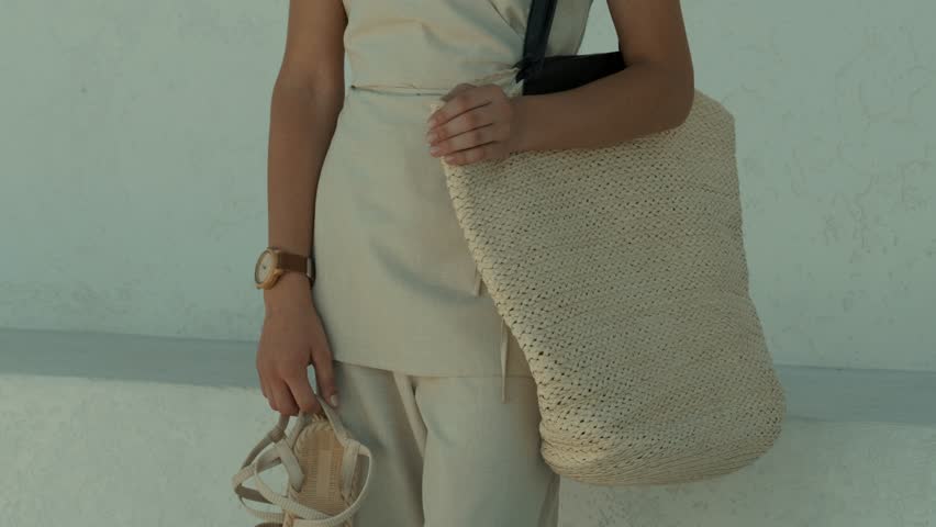 A woman wearing a neutral-toned linen ensemble walks with a spacious, textured straw tote bag slung over her shoulder, showcasing summer fashion
