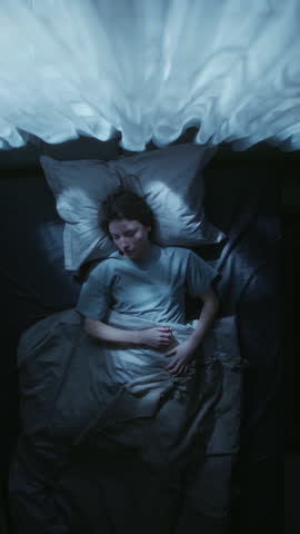 Vertical shot of young adult woman nervously rolling over in bed trying to get comfortable while struggling to fall asleep during night in bedroom