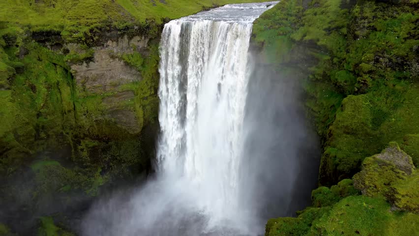 Skógafoss Waterfall in Iceland from above, drone zoom out, going back cinematic video, green grass and tourists, summer
