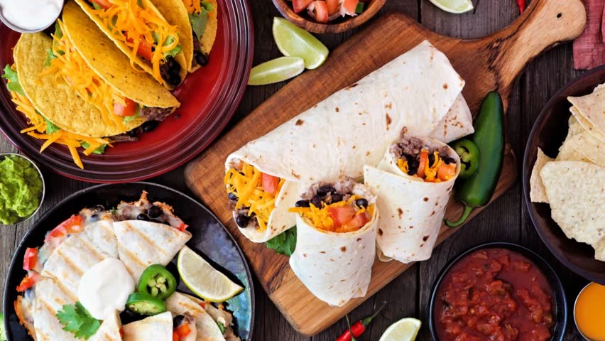 Mexican food table scene, above view on a dark wood background. Slow rotating zoom motion. Tacos, burritos, quesadilla and nachos.