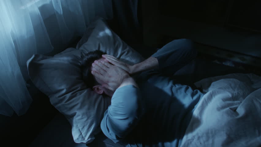 Middle aged man suffering from sleep deprivation feeling discomfort while lying in bed, tossing pillow up to get to sleep at late night