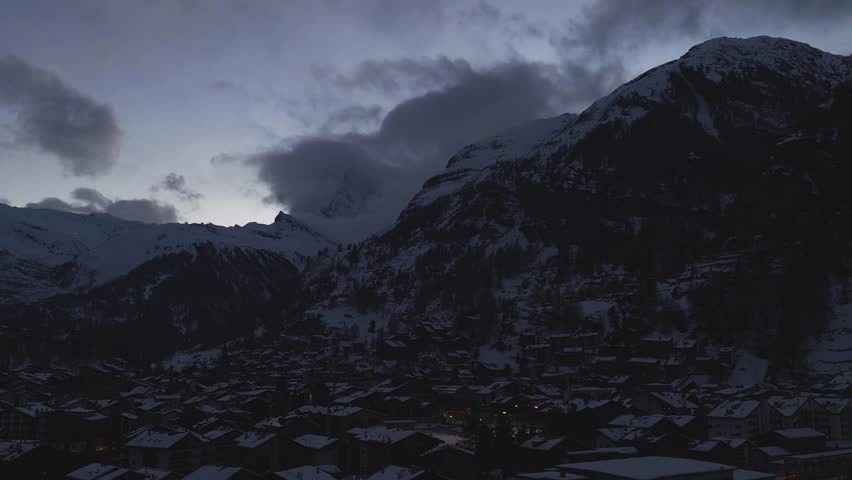 Aerial view of Zermatt in Switzerland as slow clouds veil the Matterhorn. Snow covers chalet roofs and warm street lights glow at twilight in wide cool light.