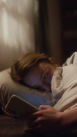 Vertical shot of young adult woman checking phone while lying in bed in morning then feeling anxiety attack building up