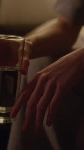 Vertical tilt up shot of young adult woman drinking from glass with quivering hands trying to soothe anxiety attack while sitting on bed in dimly lit bedroom