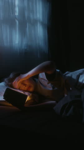 Vertical shot of young adult woman scrolling through worrying posts on phone while lying in bed during late night postponing sleep
