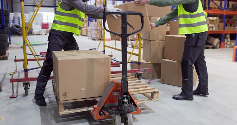 Workers loading boxes with pallet truck in warehouse. Two workers in safety vests move parcels and packages for delivery, shipping and storage. Concept of efficient warehouse logistics teamwork.