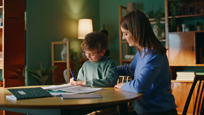 Happy mother son teaching home together enjoying teamwork closeup. Smiling young woman supporting little schoolboy in elementary school task sitting apartment workplace. Cute pupil writing notebook.