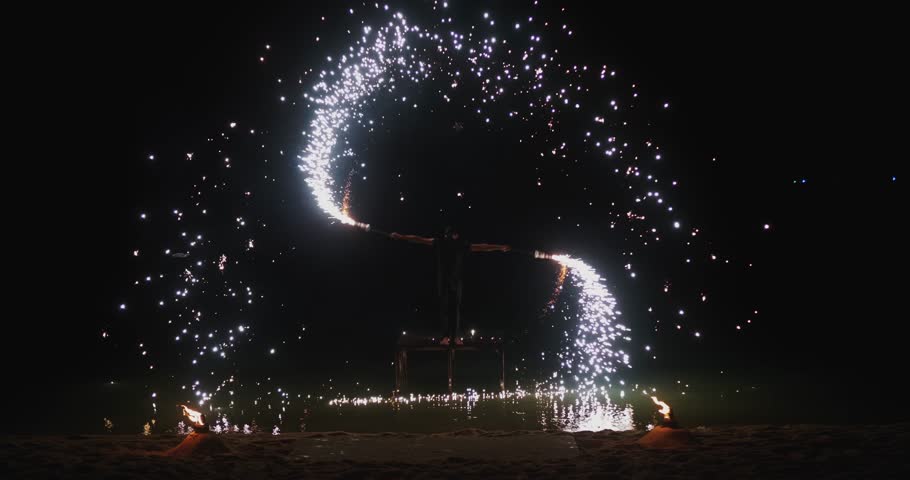 Sparks stream from burning flares as fire dancer performs during fireworks on a dark beach. Sparks and fireworks illuminate thick smoke glowing water reflections and powerful night performance energy