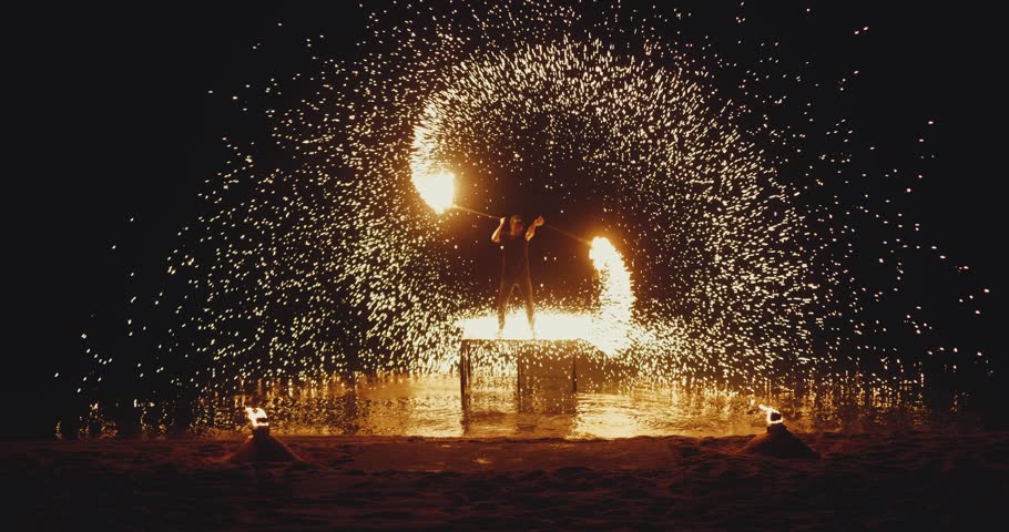 Fire dancer spins burning poi creating a massive spiral of golden sparks around body in the dark night. Fire dancer performance with glowing sparks and fire motion forms fire dancer spectacle on beach