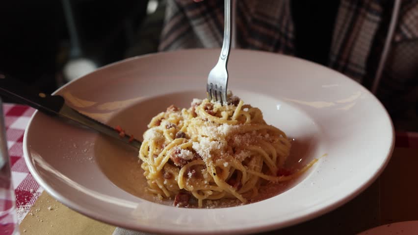 Hand holding fork twirls creamy spaghetti carbonara, topped with grated cheese and pancetta, in white bowl on red checkered tablecloth.