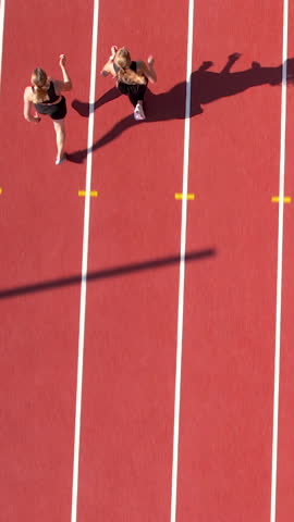 Vertical overhead drone shot of two young women competing in a sprint on a red stadium track, highlighting speed and athletic performance.