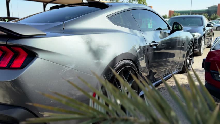 Detailed rear side view of a metallic gray performance sports coupe showing taillights, rear spoiler, alloy wheel, and sleek body lines parked outdoors in a daylight city parking lot.