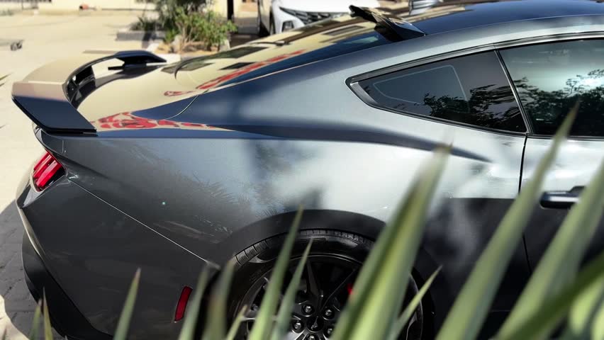 Side rear close-up of a gray performance sports coupe showing aerodynamic spoiler, rear quarter panel, and alloy wheel parked outdoors in daylight urban parking environment.